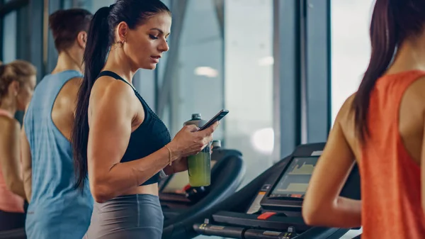 Schöne Sportlerin im Fitnessstudio, die auf einem Laufband läuft, nutzt Smartphone und trinkt Proteinsupplement Hydratationsflüssigkeit aus dem Tumbler. Im Hintergrund fitte Sportler beim Training. Seitenansicht — Stockfoto