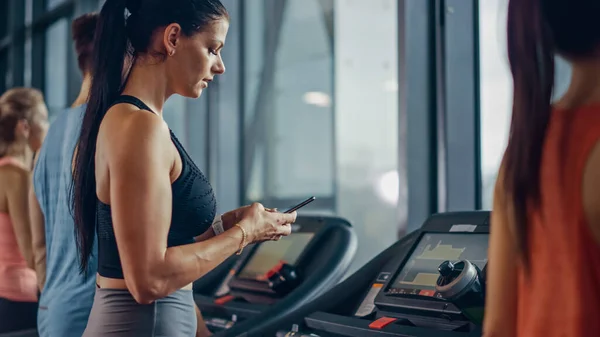 Belle sportive sportive femme dans la salle de gym Courir sur un tapis roulant, utilise Smartphone. Contexte S'adapter à l'entraînement des athlètes. Vue latérale — Photo