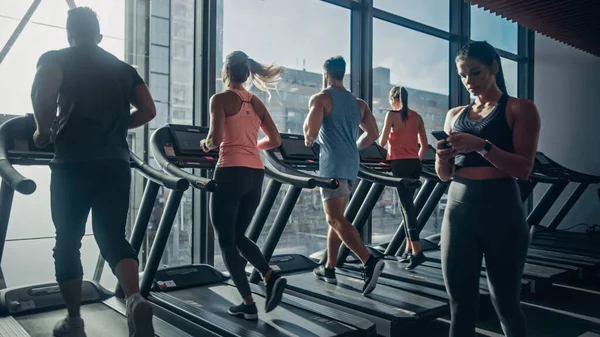 Belle femme athlétique utilise Smartphone marche à travers Fitness Club Gym. Utilisation des médias sociaux, affichage d'images, communication, vérification des courriels. En arrière-plan Sports Les gens qui courent sur les tapis roulants — Photo