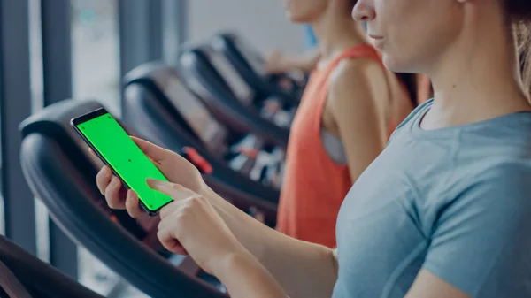 Sportowa kobieta robi gesty podczas korzystania z zielonego ekranu smartfona i bieganie na bieżni w siłowni. Fit Kobieta Posty w mediach społecznościowych, Robi zdjęcia, Ogląda filmy, Ćwiczenia w klubie fitness — Zdjęcie stockowe
