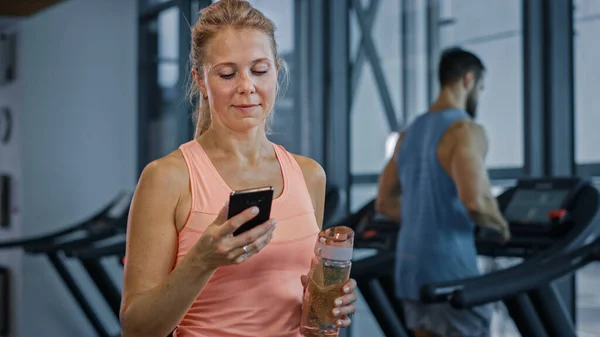 Beautiful Athletic Woman Uses Smartphone Walks Through Fitness Club Gym. Using Social Media, Posting Pictures, Communicating, Checking Email. In the Background Sports People Running on Treadmills