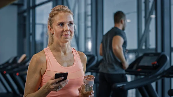 Mulher Atlética bonita usa Smartphone Walks Through Fitness Club Gym. Usando mídias sociais, postando imagens, se comunicando, verificando e-mail. No fundo Desportivo Pessoas Correndo em Treadmills — Fotografia de Stock