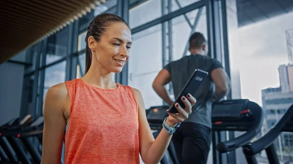 Piękna atletyczna kobieta używa smartfona Spacery przez siłownię klubu fitness. Korzystanie z mediów społecznościowych, zamieszczanie zdjęć, komunikowanie się, sprawdzanie poczty e-mail. W tle Sportowcy biegający na bieżniach — Zdjęcie stockowe
