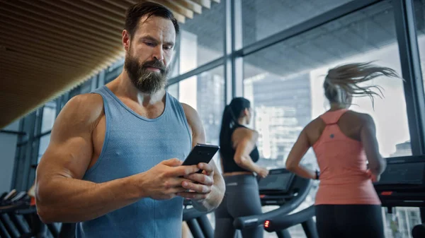 Champion musclé poids lourd se promène dans la salle de gym, utilise Smartphone pour les médias sociaux et la conduite des affaires. En arrière-plan Sports Les gens qui courent sur les tapis roulants — Photo