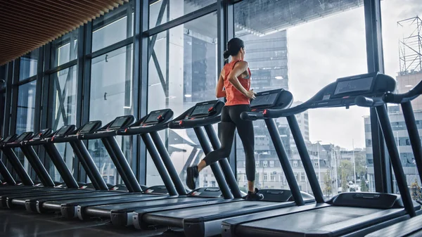 Beautiful Athletic Sports Woman Running on a Treadmill. Energetic Fit Female Athlete Training in the Gym Alone. Urban Business District Window View. Side Back View