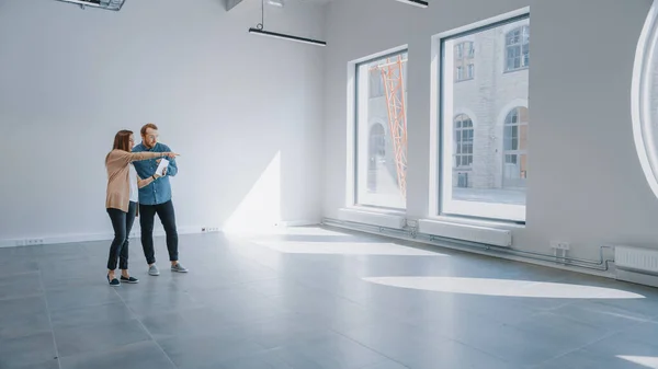 Junge Hipster stehen in einem leeren weißen Büro und kartieren es mit einer Augmented-Reality-Software auf einem Tablet. Sonnenlicht scheint durch große Fenster. — Stockfoto