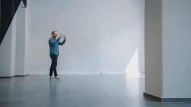 Kot Gömlekli Genç Hippi Boş Ofis 'te duruyor ve Tablet' te Artırılmış Gerçeklik Yazılımı ile haritasını çıkarıyor. Güneş ışığı Büyük Pencerelerden Parlıyor.