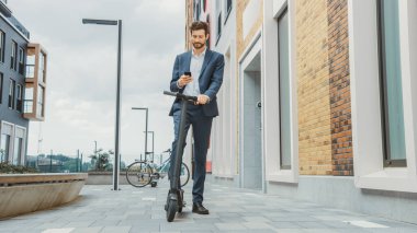 Genç İşadamı, elektrikli scooter 'la işe giderken akıllı telefonunu kontrol ediyor. Modern Girişimci Ofis Toplantısına Çağdaş Ekolojik Taşımacılık Kullanıyor.