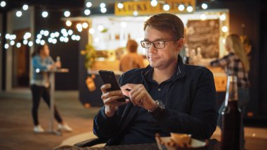 Gözlüklü Yakışıklı Genç Adam, Açık Hava Sokak Yemekleri Kafesi 'nde bir masada otururken ve kızartma yerken akıllı telefon kullanıyor. İnternet ya da Sosyal Medya Taranıyor, Durum Güncellemesi Gönderiliyor.