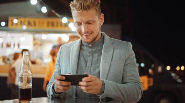 Yakışıklı Genç Adam Günlük Takımla Çok Bağımlı Online Video Oyunu Oynuyor. Outdoor Street Food Cafe 'de bir masada otururken akıllı telefon kullanıyordu. İndirdiği Uygulama.