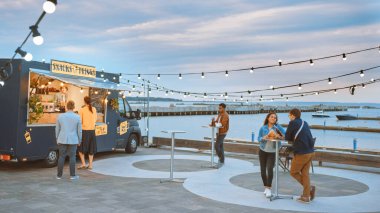 Yemek kamyonu çalışanı hamburger, patates kızartması ve soğuk içecekleri mutlu hippi müşterilere dağıtıyor. İnsanlar dışarıda masalarda yemek yiyor. Ticari Kamyon Deniz Kenarı Modern Bir Yer 'de Sokak Yiyeceği Satıyor.