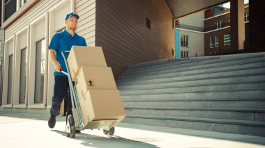 Happy Young Delivery Man Pushes Hand Truck Trolley Full of Cardboard Boxes and Packages For Delivery. Professional Courier Working Efficiently and Quickly. In the Background Stylish Modern Urban Area clipart