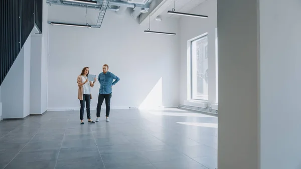 Молодой хипстер-мен и женщина стоят в пустом белом офисе и отображают его с помощью программного обеспечения дополненной реальности на планшете. Солнечные лучи сквозь большие окна . — стоковое фото