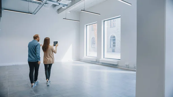 Junge Hipster stehen in einem leeren weißen Büro und kartieren es mit einer Augmented-Reality-Software auf einem Tablet. Sonnenlicht scheint durch große Fenster. — Stockfoto