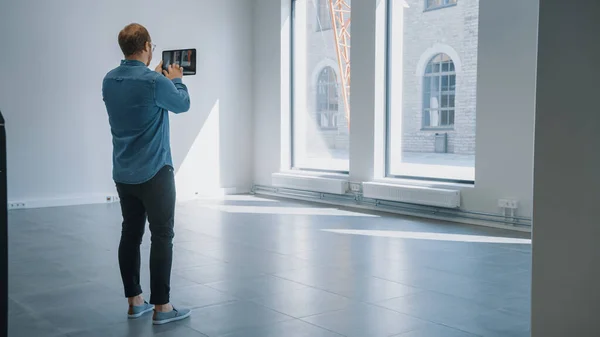 Young Hipster Man in Glasses Standing in Empty Office e mapeá-lo com um software de realidade aumentada em um tablet. Luz solar brilha através de grandes janelas . — Fotografia de Stock