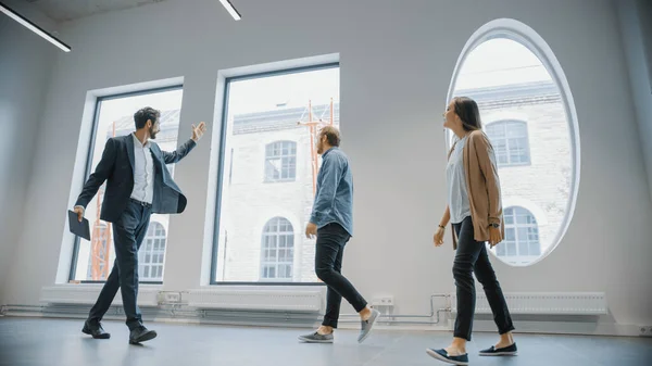 Immobilienmakler zeigt jungen männlichen und weiblichen Hipstern einen neuen leeren Büroraum. Unternehmer treffen den Makler mit einem Tablet und besprechen die Einrichtung, die sie kaufen oder mieten möchten. — Stockfoto