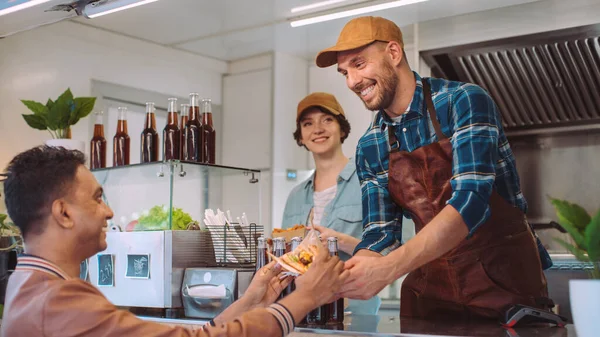 フードトラック従業員は幸せな若いヒップスターの顧客にハンバーガーと冷たい飲み物を手に入れます。現代のクールな近所の商業トラック販売ストリートフード. — ストック写真