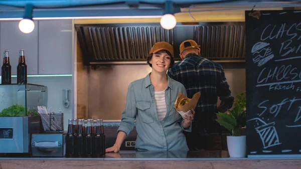 Camion de nourriture Employé féminin Tenez hamburger savoureux, sourit et regarde la caméra. Camion de nourriture de rue Vendre de la nourriture de rue dans un quartier moderne. Propriétaire d'entreprise est heureux au travail. Brunette porte un bonnet. — Photo