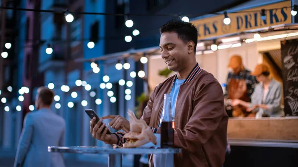 Handsome Young Indian Man is met behulp van een smartphone tijdens het zitten aan een tafel in een Outdoors Street Food Cafe. Hij surfen op het internet of sociale media, het plaatsen van een status-update. De mens is gelukkig en lachend. — Stockfoto