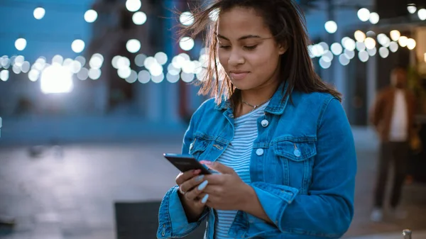 Όμορφη Μπρουνέτ Νεαρή Γυναίκα Χρησιμοποιεί Ένα Smartphone Ενώ Στέκεται Έξω Το Βράδυ. Περιήγηση στο διαδίκτυο ή στα μέσα κοινωνικής δικτύωσης, ανάρτηση ενημέρωσης κατάστασης. — Φωτογραφία Αρχείου