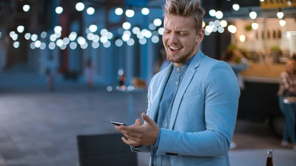 Όμορφος Happy Young Man χρησιμοποιεί ένα Smartphone, ενώ στέκεται έξω το βράδυ. Περιήγηση στο διαδίκτυο ή στα μέσα κοινωνικής δικτύωσης, ανάρτηση ενημέρωσης κατάστασης. Ο άνθρωπος φοράει κοστούμι.. — Φωτογραφία Αρχείου