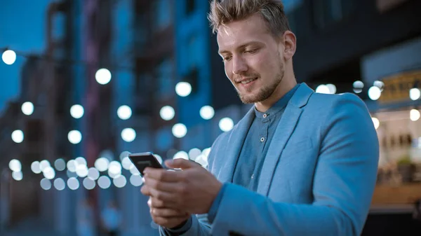 Schöner junger, konzentrierter Mann benutzt ein Smartphone, während er abends im Freien steht. Er surft im Internet oder in den sozialen Medien und postet ein Statusupdate. Mann trägt Anzug. — Stockfoto