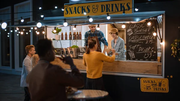 Food Truck Employee Hands Out a Freshly Made Burger to a Happy Young Female. Young Lady is Paying for Food with Contactless Credit Card. Street Food Truck Selling Burgers in a Modern Hip Neighbourhood