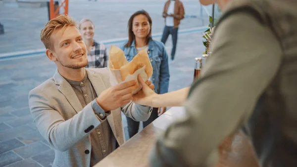 Food Truck Employee Hands Out Burger appena fatto per un felice giovane uomo in giacca e cravatta. Street Food Truck vendita hamburger in un quartiere moderno dell'anca — Foto Stock