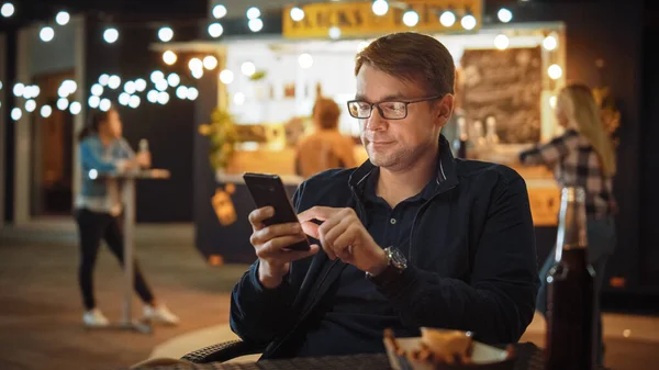 Handsome Young Man in Glasses is met behulp van een smartphone tijdens het zitten aan een tafel in een Outdoors Street Food Cafe en het eten van frietjes. Hij surfen op het internet of sociale media, het plaatsen van een status-update. — Stockfoto