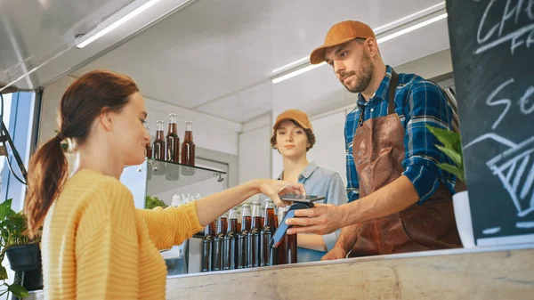 A jovem está usando o pagamento móvel da NFC para pagar por comida. Food Truck Employee Goinig para distribuir um hambúrguer recém-feito para uma fêmea. Street Food Truck vendendo hambúrgueres fora . — Fotografia de Stock