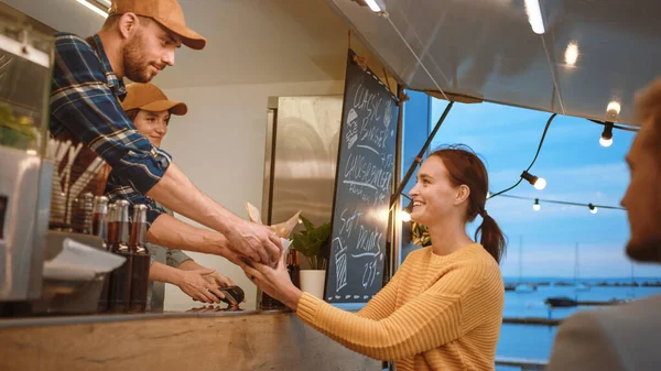 餐车员工向一位快乐的年轻女性分发新制造的汉堡。年轻的女士正在用不可接触的信用卡购买食物。现代平房街坊的街头食品卡车销售汉堡 — 图库照片