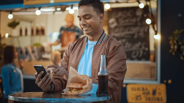 Όμορφος νεαρός Ινδός χρησιμοποιεί ένα Smartphone, ενώ κάθεται σε ένα τραπέζι σε μια υπαίθρια Cafe Τροφίμων Street. Περιήγηση στο διαδίκτυο ή στα μέσα κοινωνικής δικτύωσης, ανάρτηση ενημέρωσης κατάστασης. Ο άνθρωπος είναι ευτυχισμένος και χαμογελαστός. — Φωτογραφία Αρχείου