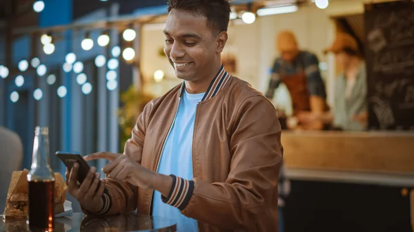 Der hübsche junge Inder sitzt mit einem Smartphone an einem Tisch in einem Street-Food-Café im Freien. Er surft im Internet oder in den sozialen Medien und postet ein Statusupdate. Der Mensch ist glücklich und lächelt. — Stockfoto