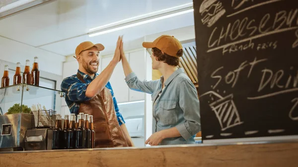 Los empleados exitosos de Food Truck, hombres y mujeres, dan los cinco primeros y celebran. Llevan gorras marrones. Son alegres y sonrientes. Camión comercial o quiosco que vende comida y bebidas callejeras . —  Fotos de Stock