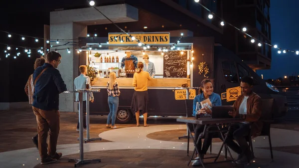 Food Truck Medewerker deelt Beef Burgers, Frietjes en Koude Drinken uit aan Happy Hipster Klanten. Mensen eten aan tafels buiten. Commerciële vrachtwagen verkoopt Street Food in een moderne plaats in de buurt van de zee. — Stockfoto