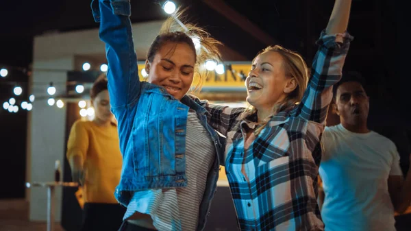 Ryhmä ystäviä pitää juhlat Street Food Burger Cafen ulkopuolella. Kaksi kaunista tyttöä halaamassa ja tanssimassa musiikin tahtiin. Ilta modernilla naapurustolla. Kaikki ovat iloisia ja iloisia.. — kuvapankkivalokuva