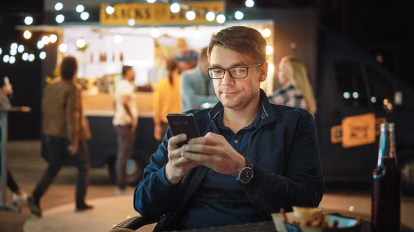 Handsome Young Man in Glasses is met behulp van een smartphone tijdens het zitten aan een tafel in een Outdoors Street Food Cafe en het eten van frietjes. Hij surfen op het internet of sociale media, het plaatsen van een status-update. — Stockfoto