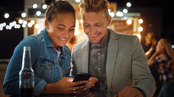 Güzel bir genç çift, bir Outdoor Street Food Cafe 'de bir masada otururken akıllı telefon kullanıyor. İnterneti ya da sosyal medyayı tarıyorlar, Videoları izliyorlar. Mutlular ve gülümsüyorlar.. — Stok fotoğraf
