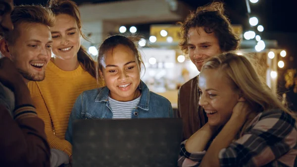 Vakre og kjekke venner bruker en laptop-datamaskin mens de står ved et bord i en utendørs gatekafe. De ser en video eller en nettstrøm. De er glade og smiler. – stockfoto