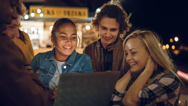 Vakre og kjekke venner bruker en laptop-datamaskin mens de står ved et bord i en utendørs gatekafe. De ser en video eller en nettstrøm. De er glade og smiler. – stockfoto