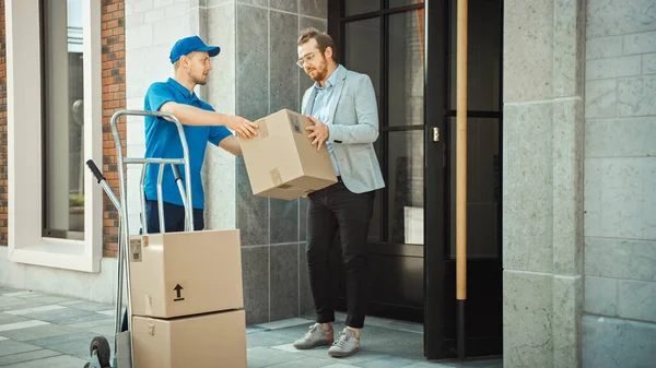 Delivery Man Pushes kézi targonca tele kartondoboz Kéz csomag egy ügyfél, aki majd aláírja az elektronikus POD eszköz. A Courier csomagokat szállít az embernek a stílusos modern városi irodaterületen — Stock Fotó