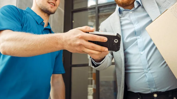 L'homme de livraison donne le paquet postal à un client d'affaires, qui signe l'appareil de PDD de signature électronique. Dans un élégant espace de bureau urbain moderne Courier livre boîte en carton colis à un homme. — Photo