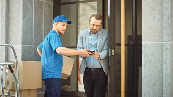 L'homme de livraison donne le paquet postal à un client d'affaires, qui signe l'appareil de PDD de signature électronique. Dans un élégant espace de bureau urbain moderne Courier livre boîte en carton colis à un homme. — Photo