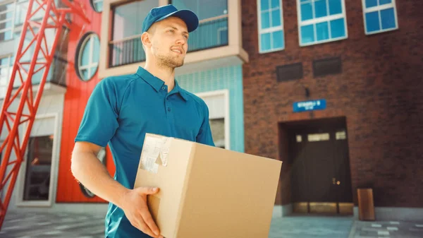 El hombre atractivo de la entrega sostiene el paquete de la caja de cartón camina a través del distrito elegante moderno del negocio. Mensajero en camino a entregar el paquete postal a un cliente . — Foto de Stock