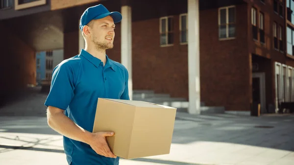 El hombre atractivo de la entrega sostiene el paquete de la caja de cartón camina a través del distrito elegante moderno del negocio. Mensajero en camino a entregar el paquete postal a un cliente . — Foto de Stock