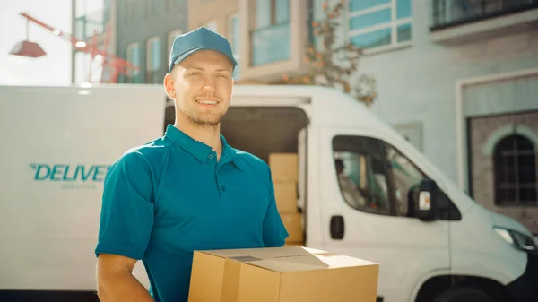 Retrato de hombre de entrega guapo sostiene paquete de caja de cartón de pie en el moderno distrito de negocios con estilo con furgoneta de entrega en segundo plano. Mensajero sonriente en camino a entregar el paquete postal al cliente — Foto de Stock