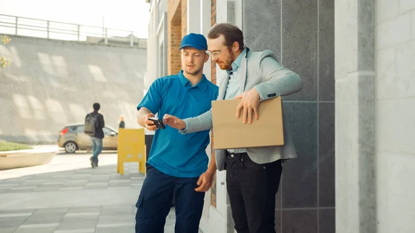 Delivery Man le da un paquete postal a un cliente comercial, que firma un dispositivo POD de firma electrónica. En la elegante oficina urbana moderna Courier entrega paquete de caja de cartón a un hombre . — Foto de Stock