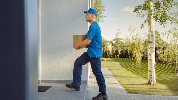 Delivery Man Holding Card Board csomag Sétál a házba. Postacsomagot kézbesítek. A háttérben gyönyörű külvárosi szomszédságában. Oldalnézet — Stock Fotó