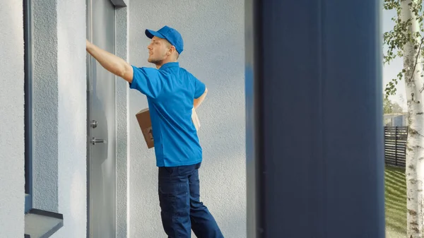 Zusteller hält Kartenbrett-Paket und klopft ans Haus. Zustellung von Postpaketen. Im Hintergrund schöne vorstädtische Nachbarschaft. Seitenansicht — Stockfoto