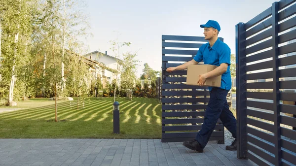 Delivery Man Holding Card Board Package gaat door de poorten en wandelingen naar het huis en klopt. Postpakketje bezorgen. In de achtergrond Prachtige buitenwijk. Zijaanzicht — Stockfoto